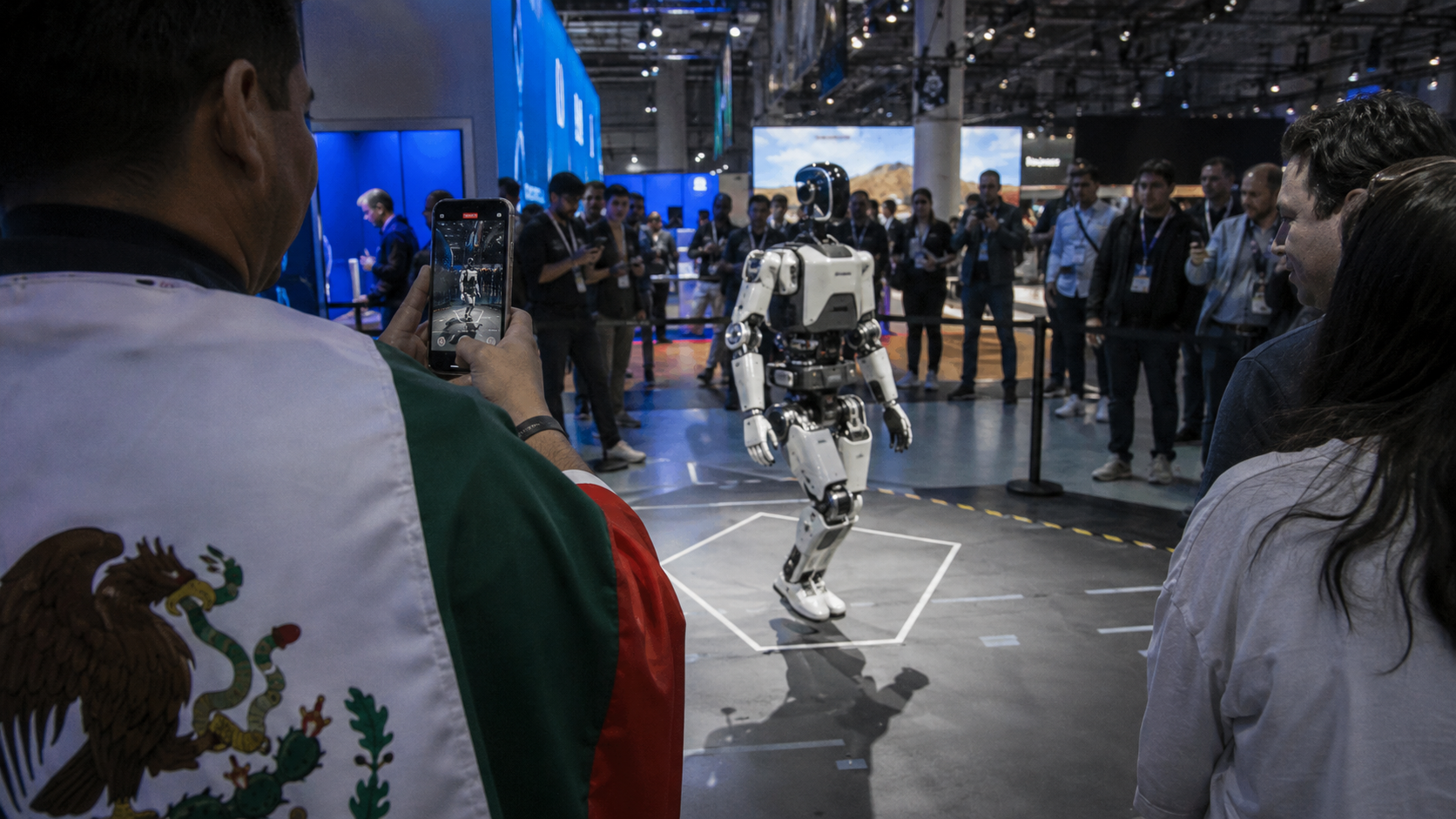 Visitante graba a un robot humanoide en una feria tecnológica, con asistentes alrededor y una persona con referencia mexicana en su ropa.